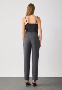 Schwarzes Satin-Camisole mit dünnen Trägern, kombiniert mit schmal geschnittenen, hochgeschnittenen anthrazitgrauen Hosen. Die Schuhe haben einen niedrigen Blockabsatz.