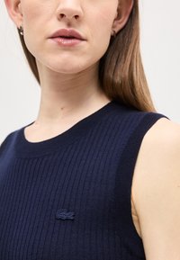 Gros plan d'une femme portant un haut sans manches côtelé bleu marine avec un petit logo crocodile brodé sur la poitrine.