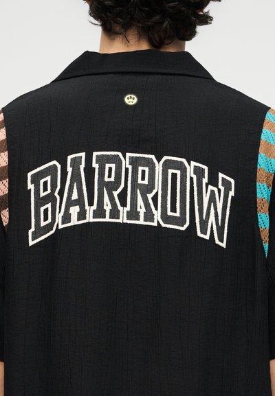 Άτομο που φοράει μαύρο μπουφάν με το κείμενο «BARROW» κεντημένο στην πλάτη και πολύχρωμα πλεκτά μανίκια ορατά κάτω από το μπουφάν.