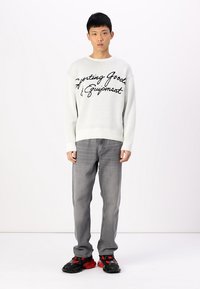 White knit sweater with "Sporting Goods & Equipment" in black script, paired with gray jeans and black sneakers with red accents.