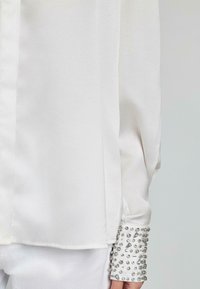 Blusa di seta bianca con una texture liscia, maniche lunghe e polsini decorati con borchie argentate.
