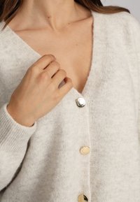 Gilet gris clair avec un col en V, des poignets côtelés et trois boutons dorés sur le devant, fabriqué en tissu doux et texturé.