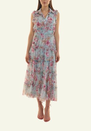 Frau trägt ärmelloses, hellblaues Blumenkleid mit gestuften Rüschen, steht mit entspannten Armen und trägt pinke Riemchenabsätze.