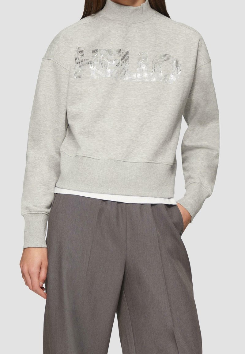 Femme portant un sweat-shirt gris clair avec "HELLO" en strass et un pantalon gris foncé ample, posant avec une main dans la poche.