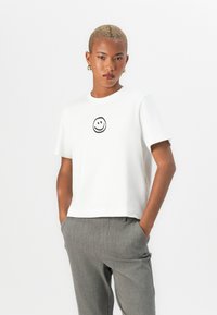 White cotton t-shirt featuring a black smiley face graphic on the front. Worn with gray textured trousers, showcasing a relaxed fit and neckline.