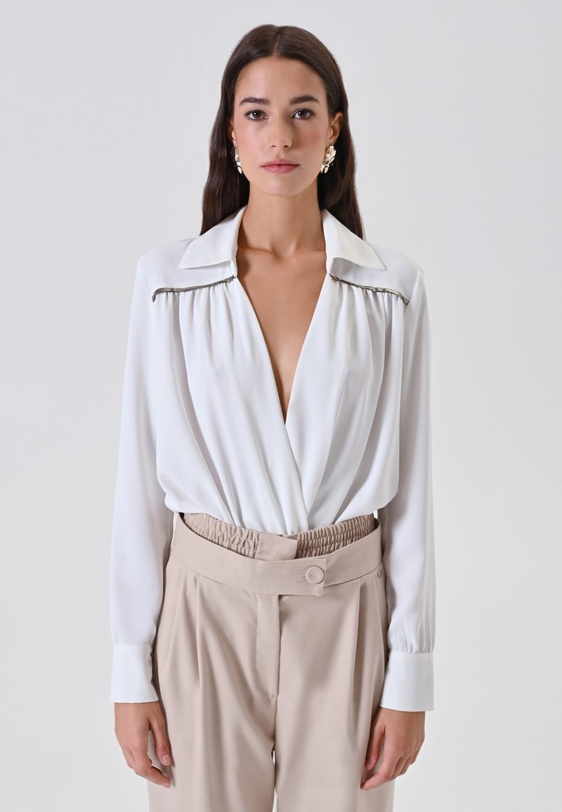 Blusa bianca a maniche lunghe, scollo a V profondo e dettagli plissettati sulle spalle, abbinata a pantaloni beige a vita alta con bottoni.