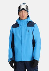 Blaue Skijacke mit marineblauen Akzenten, ausgestattet mit einem hohen Kragen, verstellbarer Kapuze und Reißverschlusstaschen. Getragen mit schwarzen Handschuhen und einer Skibrille.