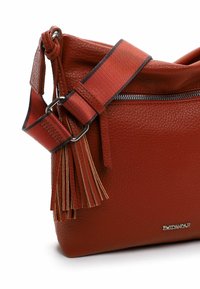Sac bandoulière en cuir rouge avec une finition texturée, une bandoulière contrastante et des franges décoratives. Dispose d'un compartiment zippé et d'un logo métallisé.