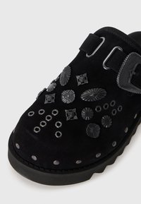 Clog din suede negru cu decorațiuni din metal negru, ochiuri și o cataramă mare pe lateral, pe un fundal alb.