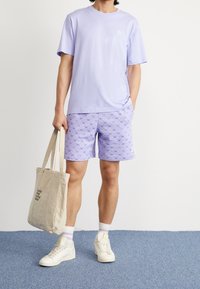 Světle fialové bavlněné tričko s krátkým rukávem a malým logem, spárované s vzorovanými fialovými šortkami a béžovou taškou. Nohou má bílé tenisky.