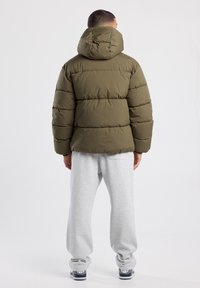 Olive green puffer jacket with a hood, featuring horizontal quilted sections and a slightly tapered waist. Worn with gray sweatpants.