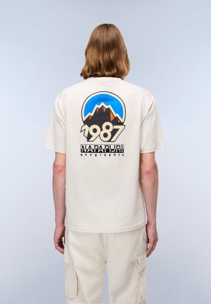 Person med skulderlangt hår bærer en lysebeige skjorte med et bjergmotiv, "1987," og "Napapijri geographic" på ryggen.