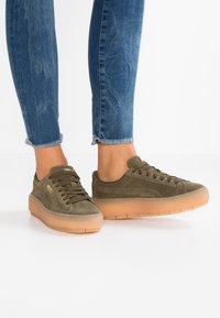Olivgrüne Wildledersneaker mit braunen Gummisohlen, ausgestattet mit runden Schnürsenkeln und einem Low-Top-Design, kombiniert mit blauen, ausgefransten Jeans.