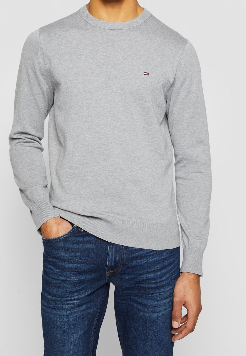 Uomo che indossa un maglione a manica lunga grigio chiaro con un piccolo logo rosso, bianco e blu sul petto, abbinato a un paio di jeans blu scuro.