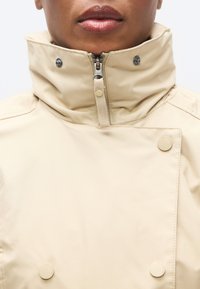 Μπεζ αδιάβροχο μπουφάν με ψηλό γιακά, εμπρόσθιο φερμουάρ και κουμπιά με κλείσιμο με πατιλέτα, με λεία, ματ υφή και κομψό σχεδιασμό.