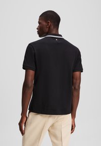 Černé polo tričko s krátkými rukávy, s bílým proužkovaným límečkem. Podrobné prošívání a malá emblematická nášivka blízko krku, bavlněný materiál.