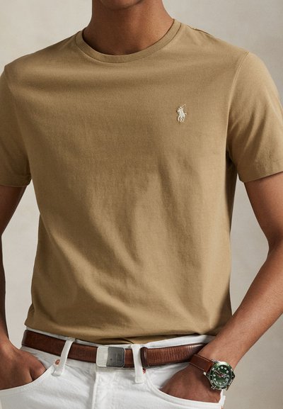 Moški, ki nosi kratko majico barve tan z belim vezenim logotipom, bele hlače z rjavim pasom in rjavo usnjeno uro.