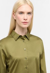 Blusa di seta verde oliva con colletto classico, dotata di una chiusura frontale a bottoni e una texture liscia. Nessun motivo o decorazione visibile.