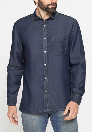 Uomo con barba che indossa una camicia di jeans blu scuro con bottoni e jeans blu chiaro, in piedi davanti a uno sfondo bianco.