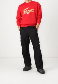 Sudadera roja con logo impreso en amarillo, pantalones cargo negros con bolsillos laterales y zapatillas negras con textura. Atuendo simple y casual.