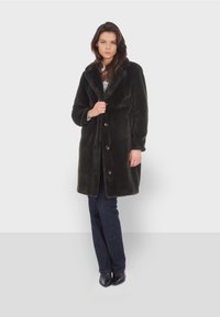 Manteau en fausse fourrure vert foncé avec un large col, des boutons à un seul boutonnage et deux poches avant, associé à un jean foncé et des talons.