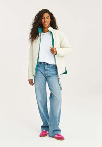 Prešívaná krémová bunda s teal podšívkou, nosená cez biele tričko. Skombinovaná s modrými džínsy a ružovými teniskami. Má zipsové zapínanie.