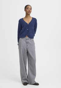 Marineblå strikket cardigan med bølgede kanter og fem knapper, parret med højtaljede, vide grå denim jeans og mørke sko.