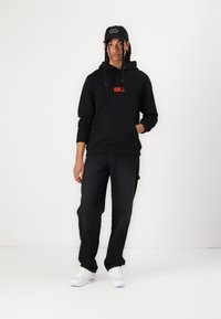 Black hoodie with red logo, paired with dark jeans and white sneakers. A black cap with text completes the outfit. Casual urban design.