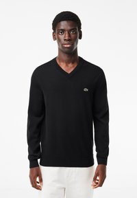 Maglione nero a scollo a V realizzato in tessuto morbido, con colletto e polsini a coste, impreziosito da un piccolo logo verde Lacoste sul petto.