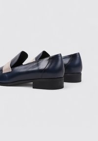 Mocasines de cuero navy con un acento en forma de V beige en contraste, tacón bajo negro y textura suave, con un diseño de fácil puesta.
