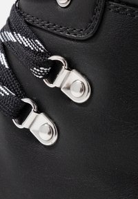 Černá kožená boots s texturovaným povrchem, černobílými pruhovanými tkaničkami a stříbrnými kovovými očky. Žádné viditelné logo ani vzory.