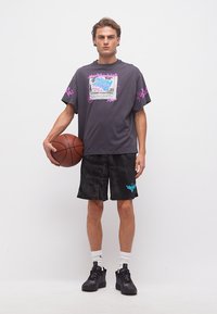 Νέος άνδρας που κρατάει μπασκετόσφαιρα, φοράει σκούρο μπλουζάκι με σχέδιο, μαύρο αθλητικό σορτς, λευκές κάλτσες και μαύρα αθλητικά παπούτσια, στέκεται μπροστά σε λευκό φόντο.