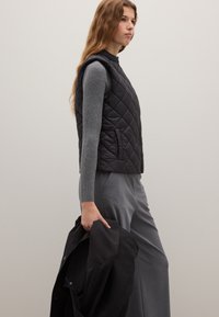 Schwarze gesteppte Weste mit hohem Kragen, kombiniert mit einem grauen Langarmshirt und weiten grauen Hosen. Eine schwarze Jacke haltend.