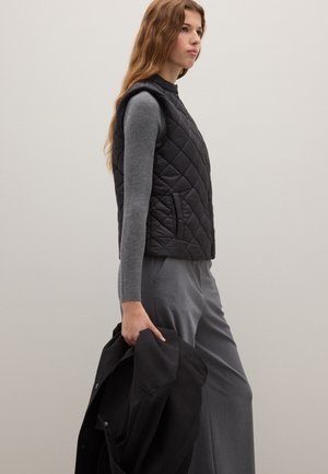 Zwarte, doorgestikte bodywarmer met een hoge hals, gecombineerd met een grijs shirt met lange mouwen en grijze wijde broek. Houdt een zwart jack vast.