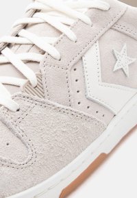 Κοντινό πλάνο ενός μπεζ σαμουέ αθλητικού παπουτσιού με λευκά κορδόνια, λευκό αστέρι και λογότυπο «βελάκι» και χείλος σόλας από καουτσούκ σε χρώμα καραμέλας.