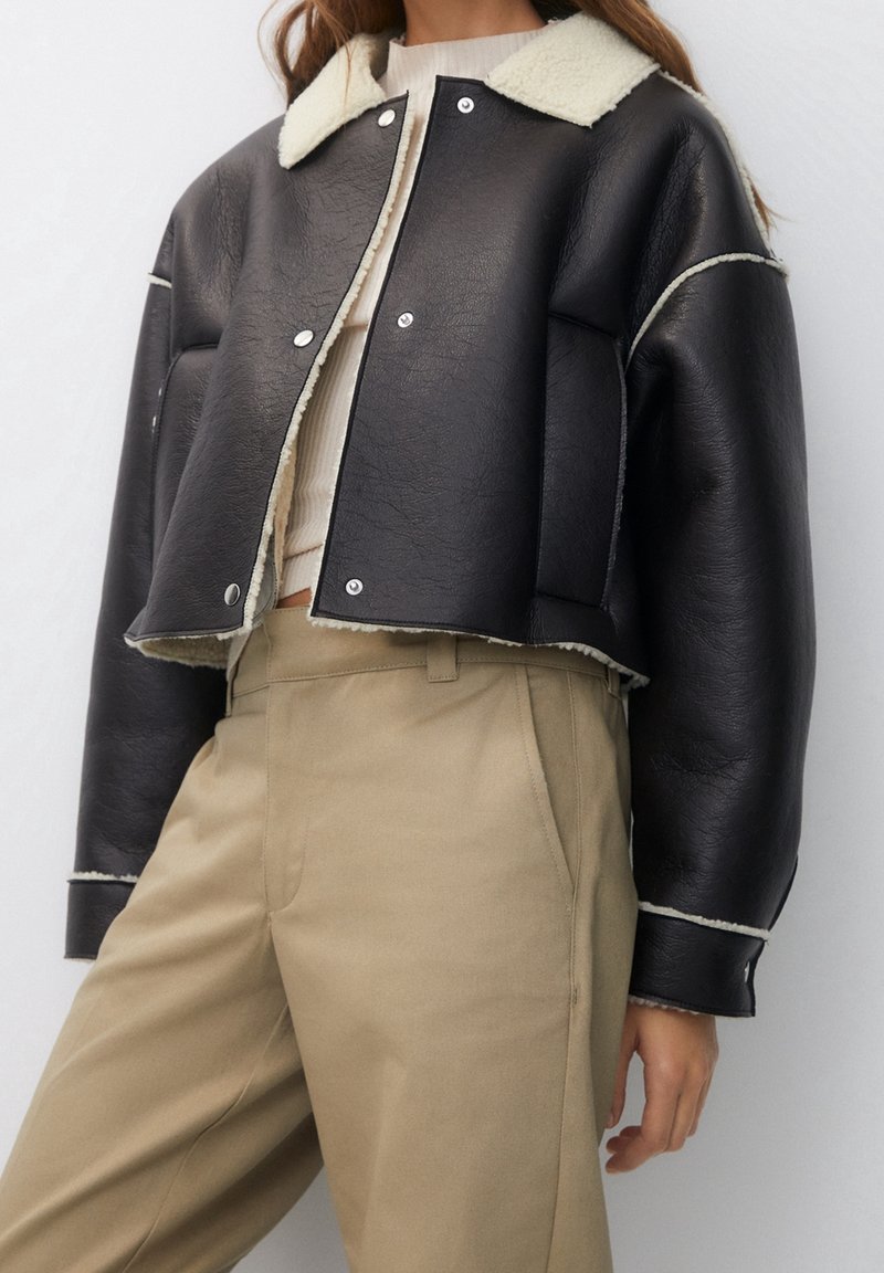 Femme portant une veste courte en simili cuir noire avec bordure en peau lainée blanche et un pantalon beige taille haute, debout devant un fond blanc uni.