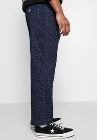 Námořnické kalhoty s leopardím potiskem a rovným střihem, se zřetelným šitím a bočními kapsami, spárované s černými nízkými teniskami.