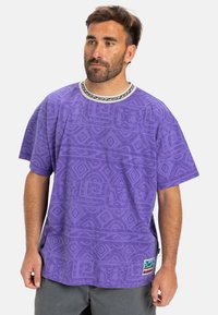 Fialová krátka rukávová tričko s textúrovaným tribal vzorom, kontrastný čierny a biely golier a nášivka s logom na leme. Voľný strih.