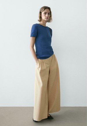 Frau trägt ein blaues Kurzarmshirt und beige Hosen mit weitem Bein, steht mit den Händen in den Taschen vor einem schlichten hellen Hintergrund.