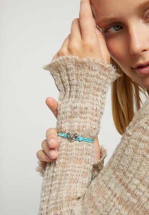 Mujer vistiendo un suéter de punto beige con mangas largas, mostrando una pulsera de cordón azul con un charm de corazón plateado en la muñeca.