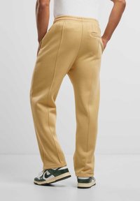 Tan joggers met een gladde textuur, een elastische tailleband, zijzakken en contrasterende stiksels, gedragen met groene en witte sneakers.