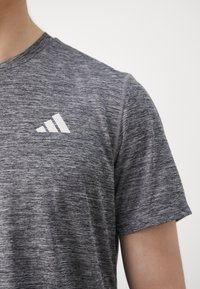Grå atletisk t-skjorte laget av lett, teksturert stoff. Har korte ermer og en hvit Adidas-logo på venstre brystområde.