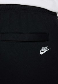 Pantalones de chándal Nike negros en primer plano, con un bolsillo trasero de apertura horizontal y un logo de Nike blanco en la parte inferior de la pierna. Textura de tela suave.
