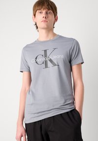 Gray cotton t-shirt featuring a large black "CK" logo with "Calvin Klein Jeans" text, crew neck, and short sleeves. Smooth texture.