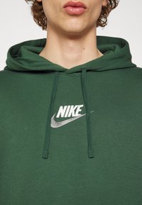 Grön bomullshoodie med dragsko i huvan; har en vit broderad Nike-logga på bröstet mot en texturerad tygbakgrund.