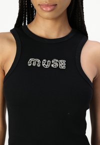 Crna rebrasta majica bez rukava s aplikacijom od rhinestona koja ispisuje "muse" u različitim veličinama i teksturama na prednjoj strani.