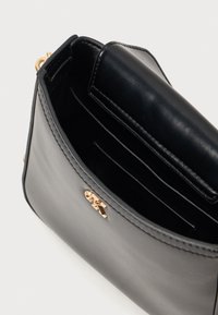 Čierna kožená kabelka s hladkou textúrou, s guľatou zlatou logovou akcentáciou a vnútrom s viacerými priehradkami na karty a zipsovým vreckom.