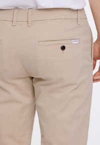 Bēša krāsas bikses ar gludu tekstūru, aizmugurējā kabatā melns pogu un maza balta etiķete uz kabatas šuves.