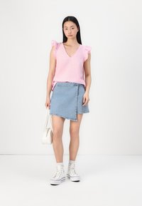 Top rosa senza maniche con spalle arricciate; design a V. Mini gonna in denim blu chiaro con taglio asimmetrico. Sneakers bianche e borsetta piccola.