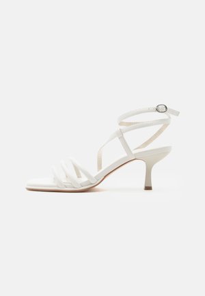 Hvid damesandal med tynde hæle, tre stropper foran og en ankelrem med en sølvspænde.